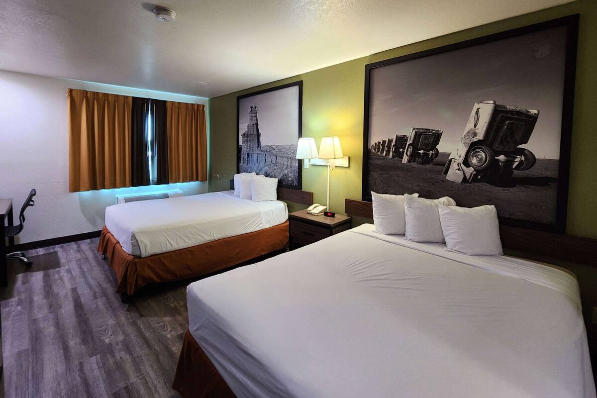 Ein Hotelzimmer mit zwei Betten, einem Schreibtisch, einer Lampe und einem großen Bild im Rahmen an der Wand.