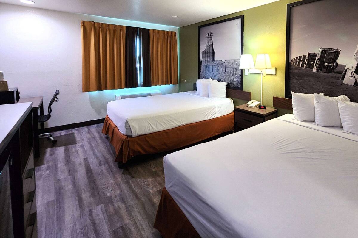 Ein Hotelzimmer mit zwei Betten, einem Schreibtisch, einem Stuhl, einer Lampe und einem gerahmten Bild an der Wand.