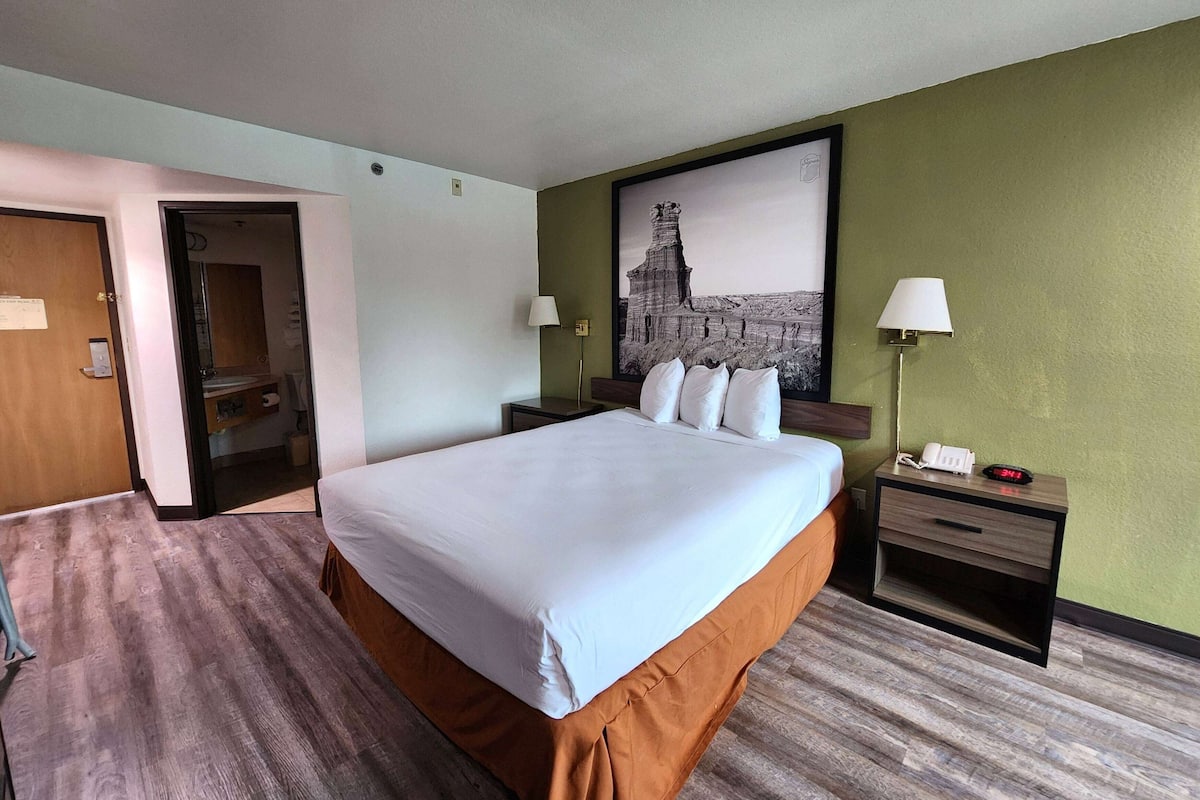 Ein Hotelzimmer mit einem großen Bett, einem Nachttisch mit Lampe, einem Bild an der Wand und einer Tür zu einem anderen Raum.