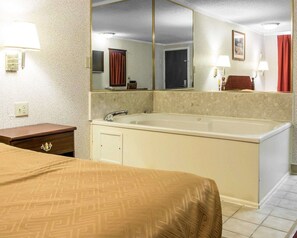 A hotel room with a bed, a large mirror, a bathtub, a nightstand, and a wall-mounted lamp. - Rodeway Inn Madison Heights - Detroit (Madison Heights)