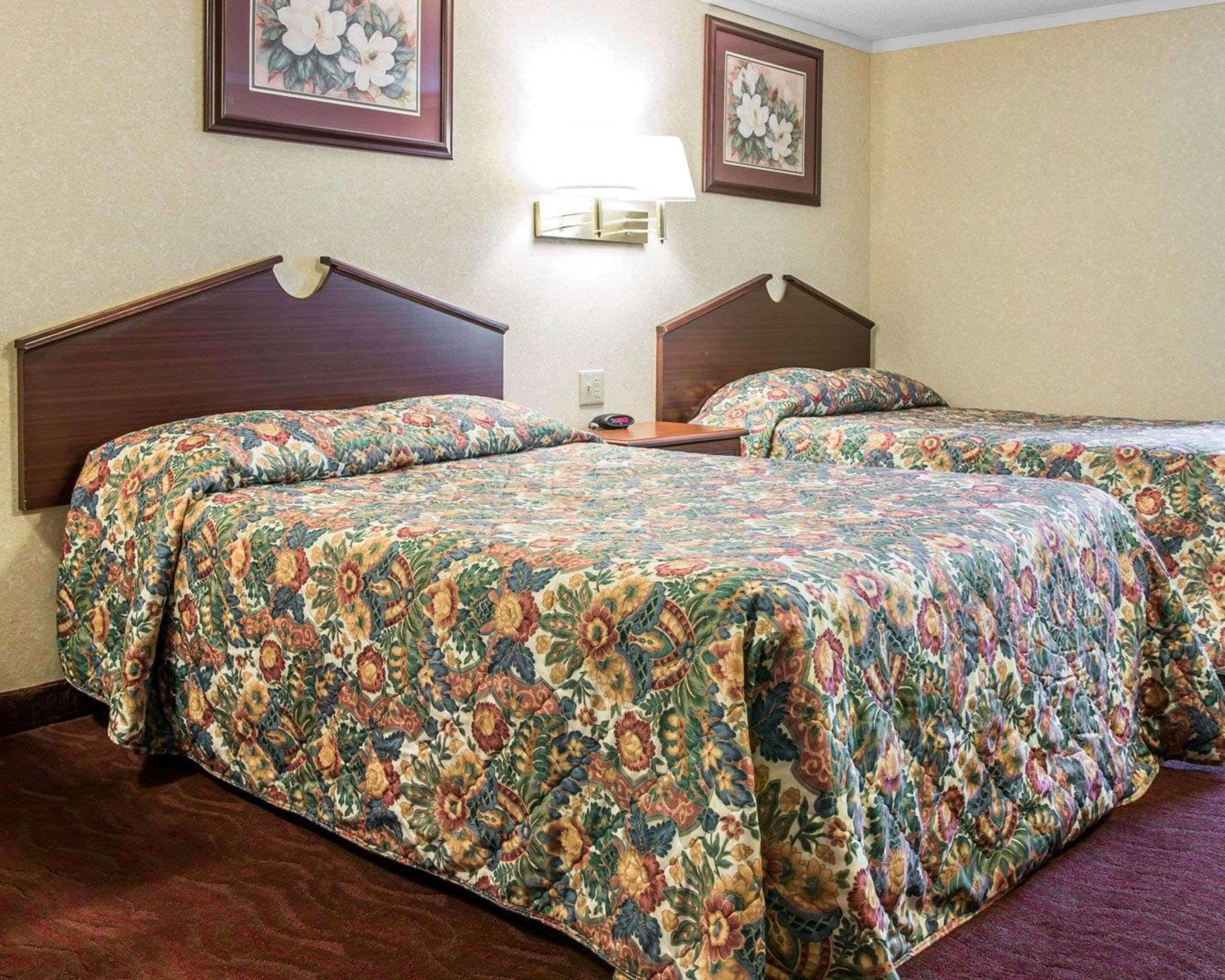 A hotel room with two beds, floral-patterned bedding, a nightstand, a lamp, and framed floral artwork on the walls.