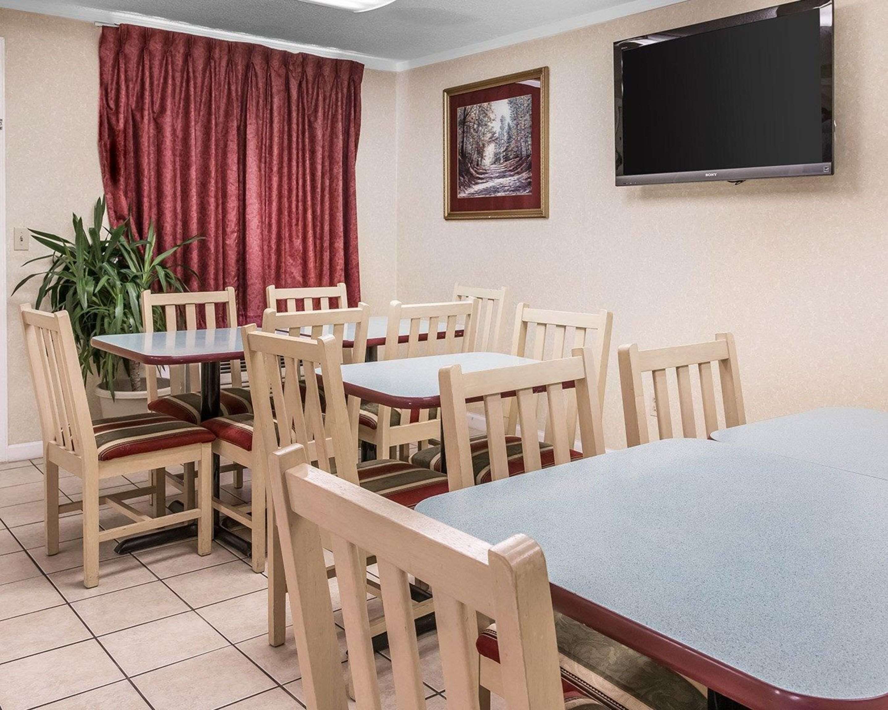 A dining area with wooden chairs and tables, a potted plant, a red curtain, a framed picture, and a flat-screen TV on the wall.