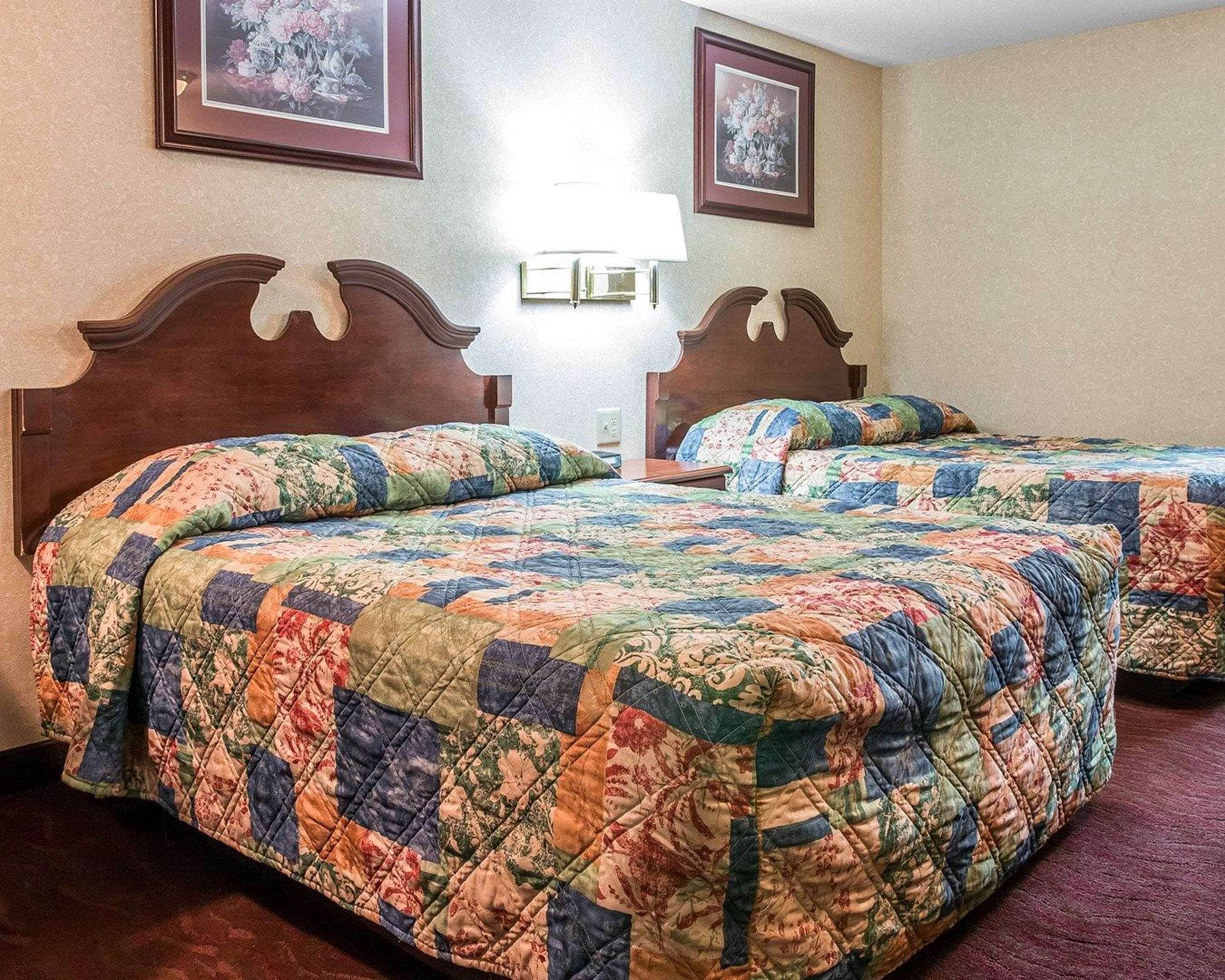 A hotel room with two beds, each with a floral quilted bedspread. There are two framed floral paintings on the wall, a bedside lamp, and a nightstand with a lamp.