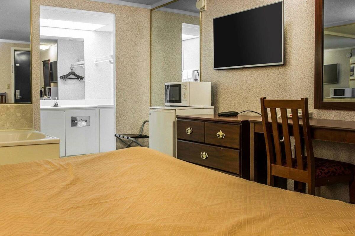 A hotel room with a bed, a wooden chair, a desk with a flat-screen TV, a microwave, a small dresser, and a mirror.