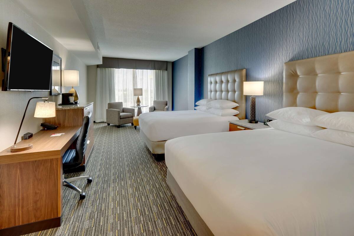 A hotel room with two beds, a desk, a chair, a TV, and a small table with lamps.