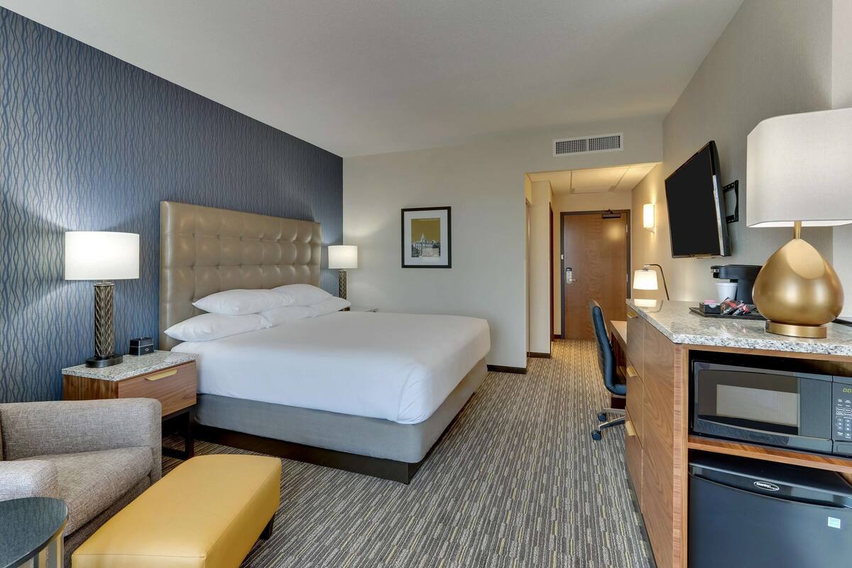 A hotel room with a bed, bedside tables, a desk with a lamp, a chair, a microwave, and a television.