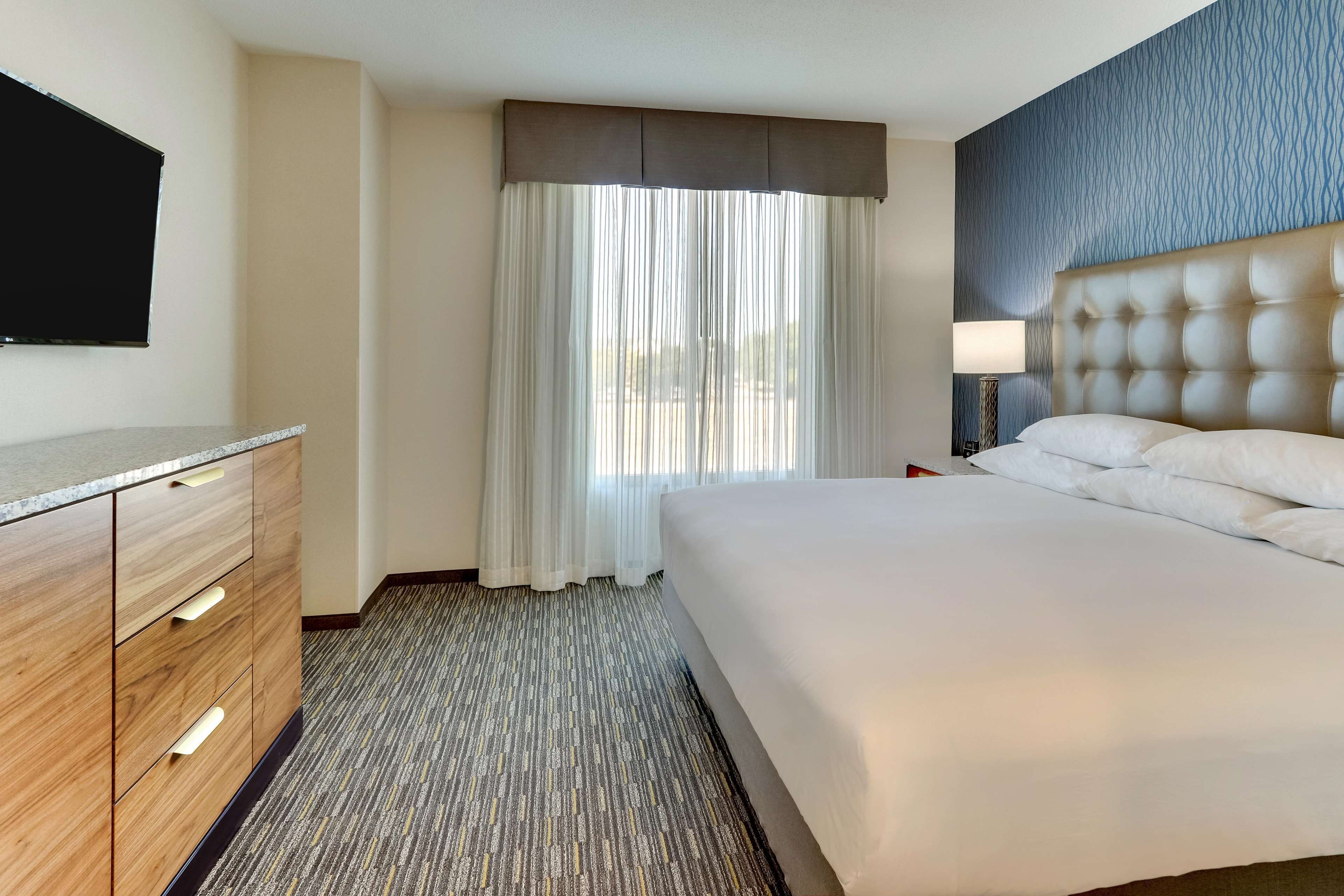 A hotel room with a large bed, a wooden dresser, a flat-screen TV, and a window with sheer curtains.