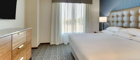 A hotel room with a large bed, a wooden dresser, a flat-screen TV, and a window with sheer curtains.