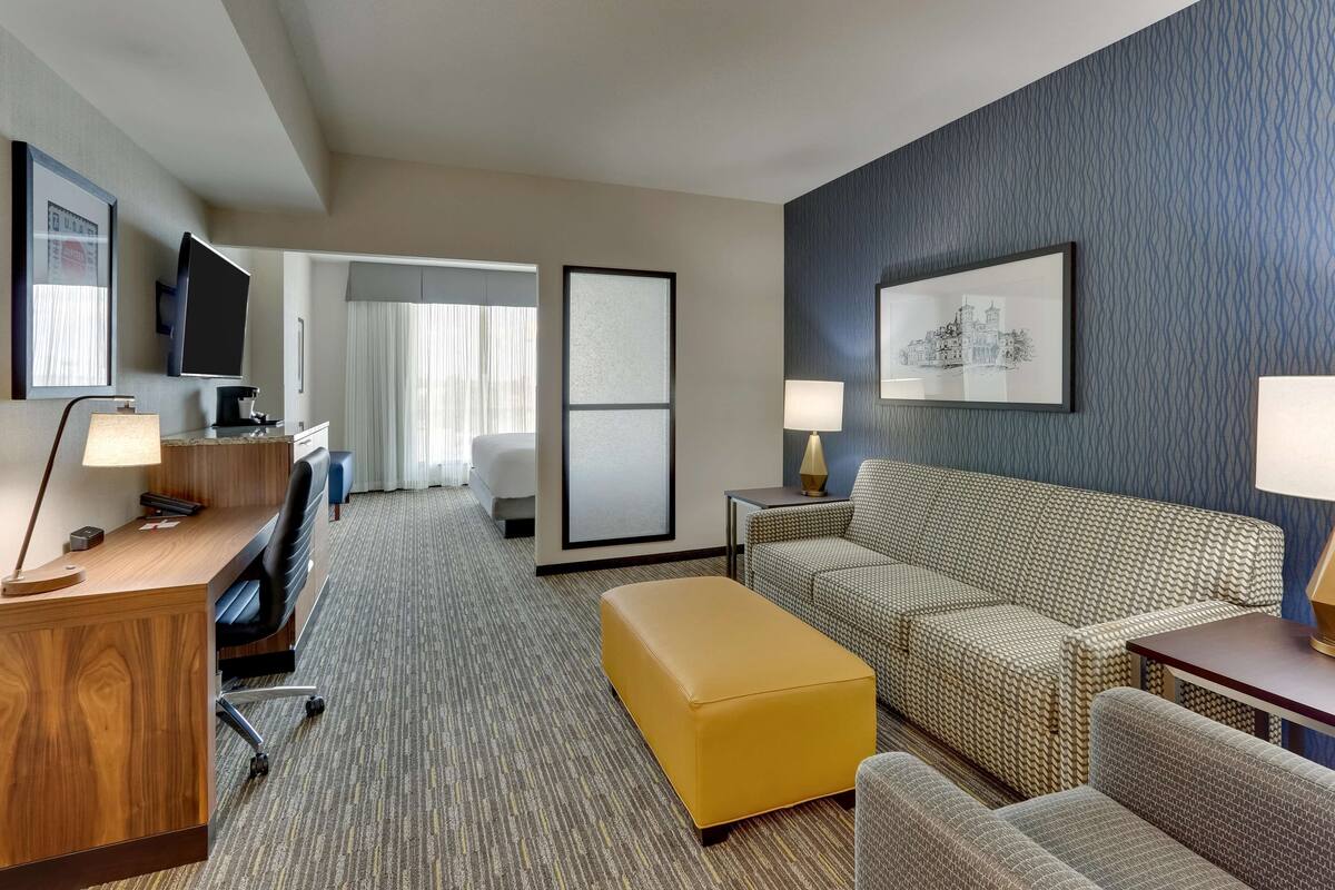 A hotel room with a wooden desk, a television, a sofa, an ottoman, and a bed.