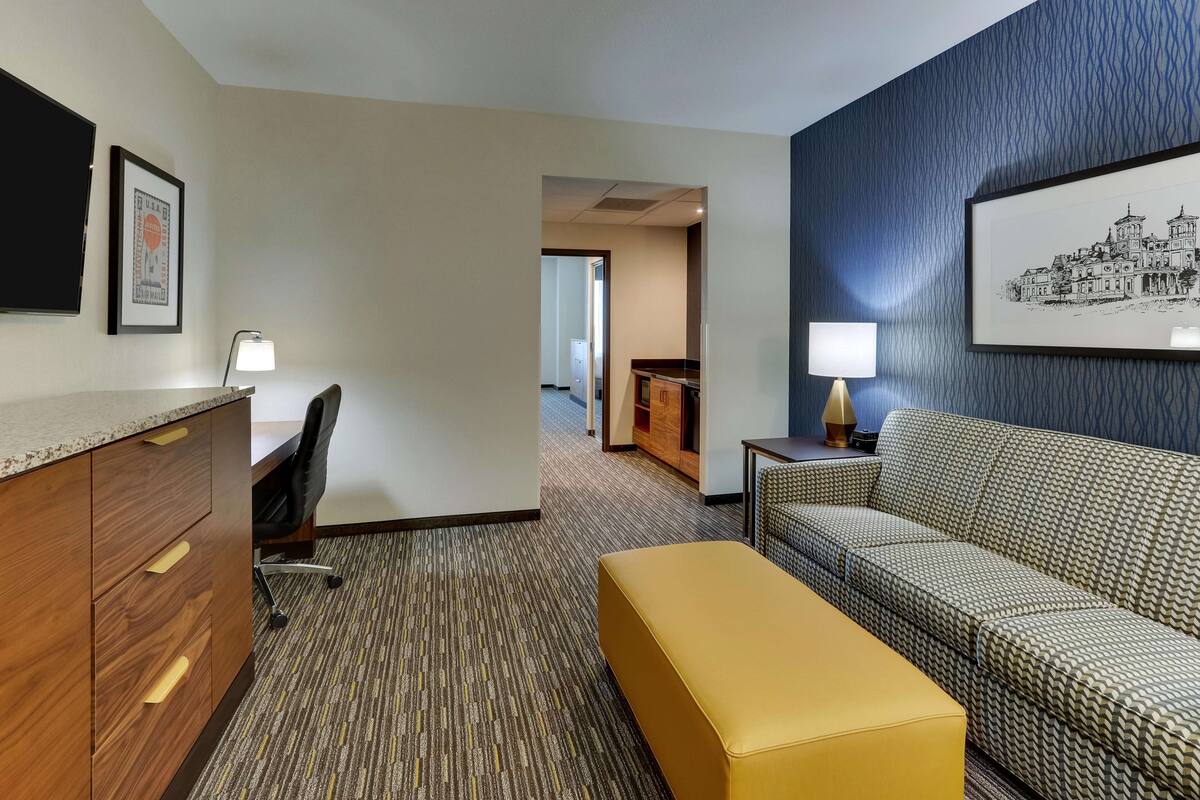 A hotel room with a wooden desk, a chair, a sofa, and a small table with a lamp.