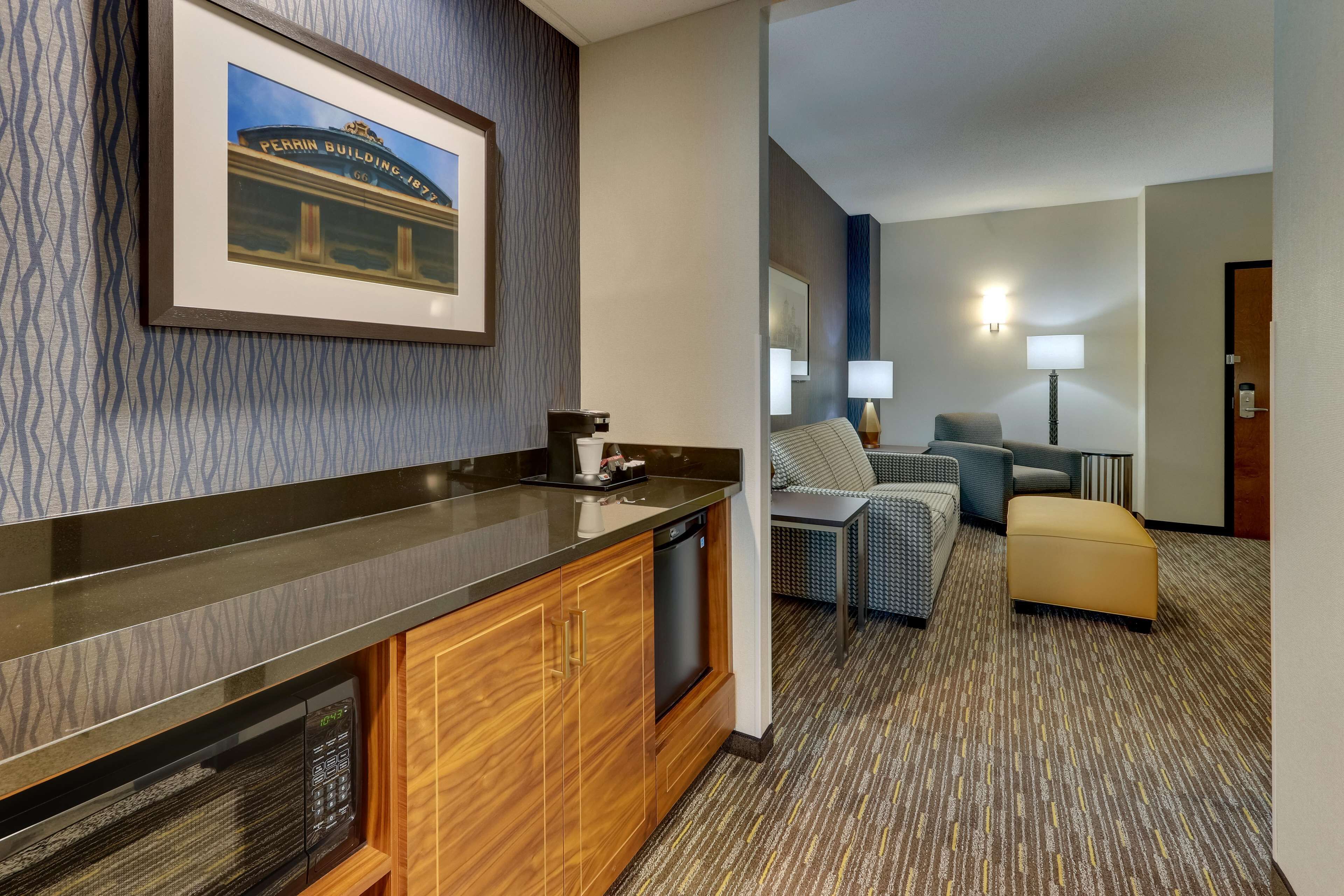 Habitación de hotel con kitchenette, un cuadro con la imagen del Edificio Perrin, un sofá, una silla y una lámpara.