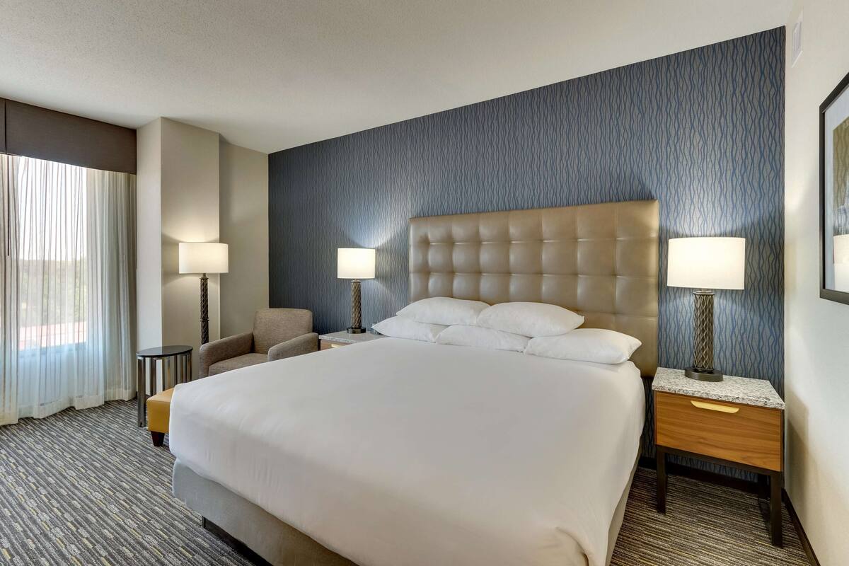 A hotel room with a large bed, two bedside tables with lamps, a chair, and a window with curtains.