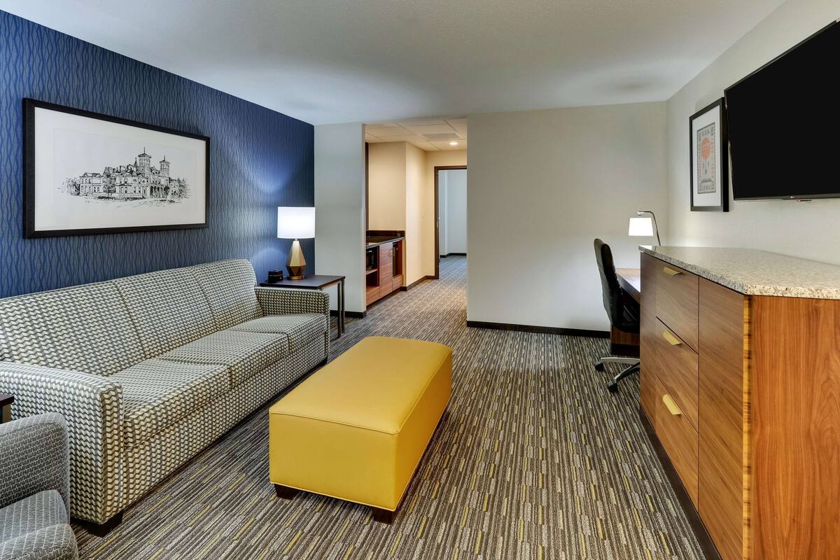 A hotel room with a patterned sofa, a yellow ottoman, a wooden desk, and a framed picture on the wall.