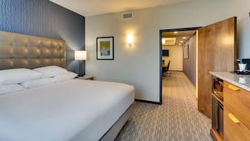 A hotel room with a large bed, a wooden headboard, a nightstand with a lamp, a framed picture on the wall, and a kitchenette area.