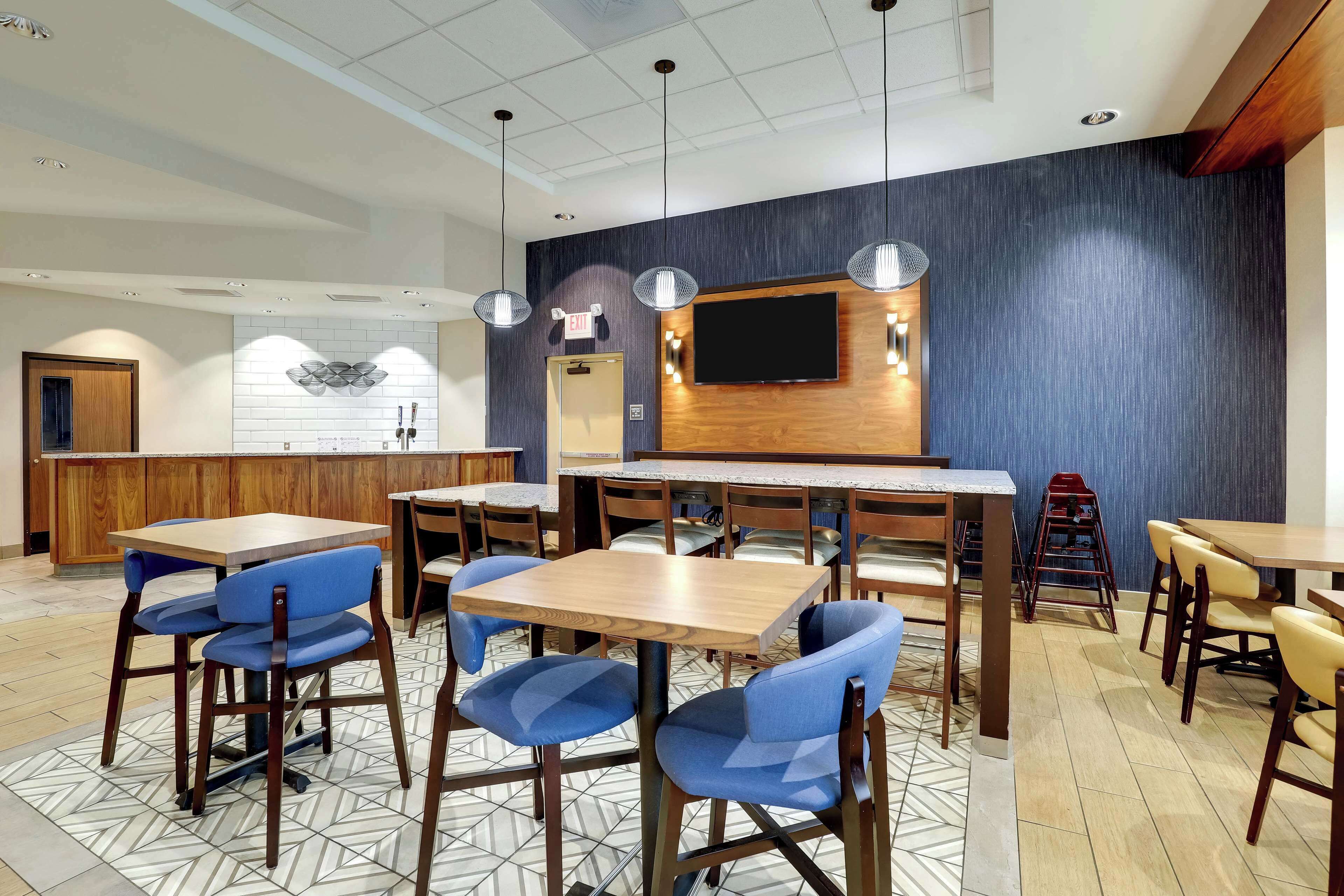 A modern restaurant with wooden tables and blue chairs, a TV mounted on the wall, and a kitchen area in the background.