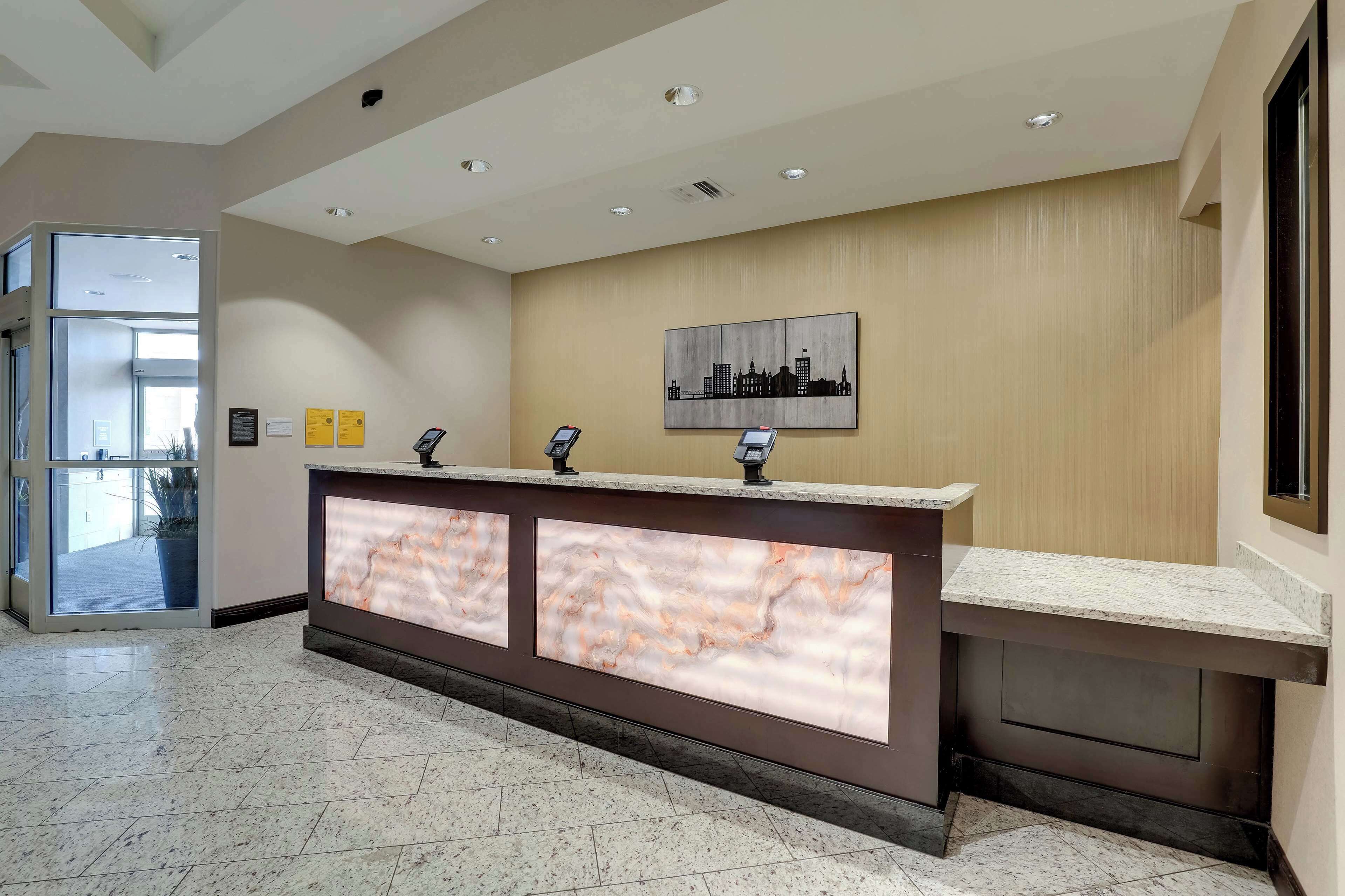 A hotel reception area with a marble floor, a reception desk with a cityscape artwork, and a glass door entrance.