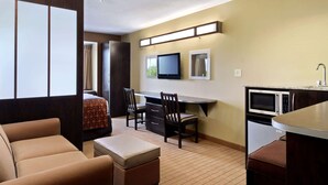 Een compacte hotelkamer met een kitchenette, een bed met een beddeken met patroon, een zithoek met een bank en stoelen, en een aan de muur bevestigde televisie.