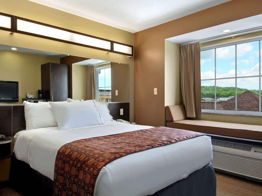 A hotel room with a large bed, a TV, a desk with a computer, a window with curtains, and a view of greenery outside.