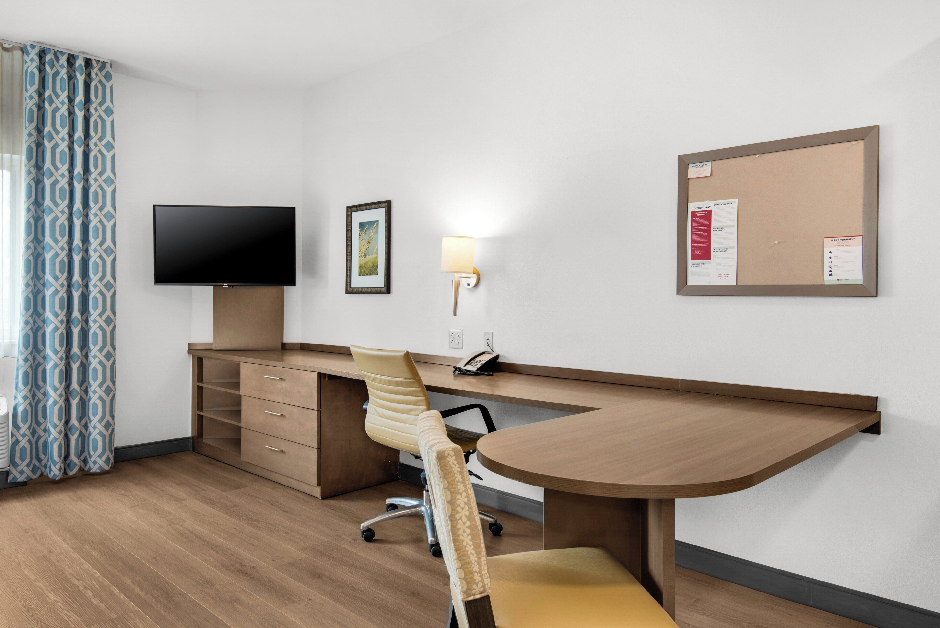 Habitación de hotel con un escritorio de madera, un televisor de pantalla plana, un sillón acolchado, una lámpara de pared y un tablón de anuncios.