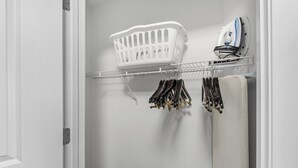 Een kast met een kledingroede, een witte wasmand en een strijkijzer op een plank.