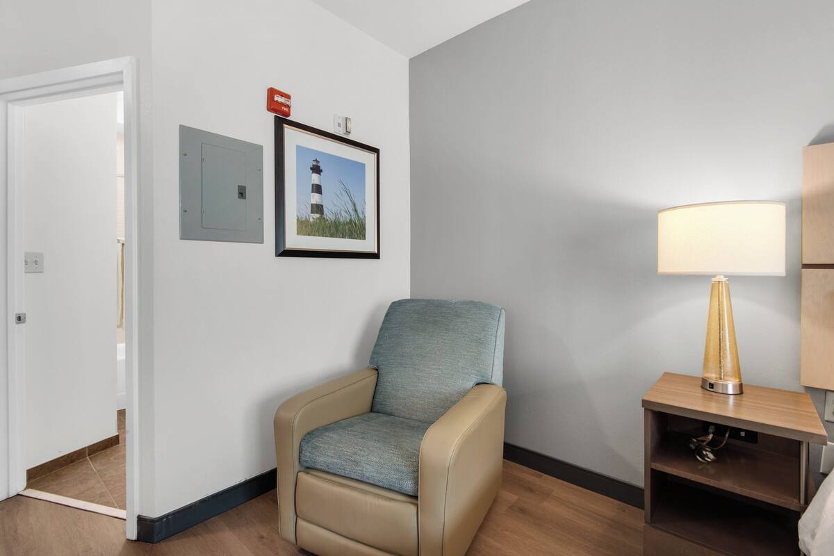Ein Hotelzimmer mit beigem Sessel, einem gerahmten Bild eines Leuchtturms, einer Nachttischlampe und einem hölzernen Nachttisch.