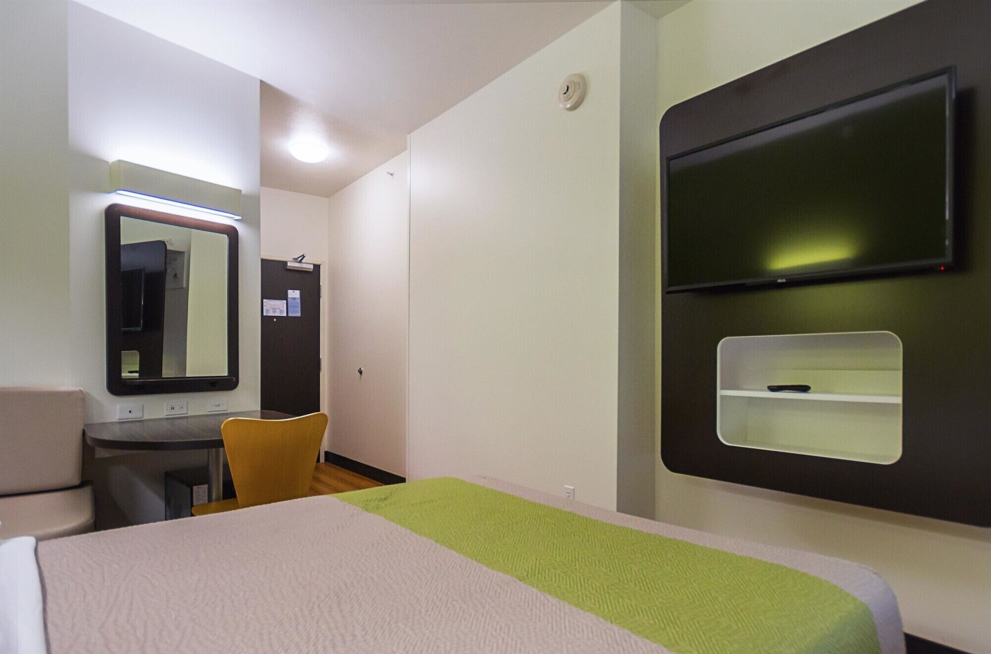 Habitación de hotel con cama, escritorio, silla, espejo, televisor y una pequeña estantería.