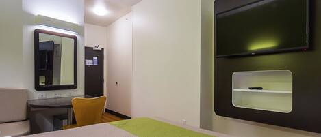 A hotel room with a bed, a desk, a chair, a mirror, a television, and a small shelf.