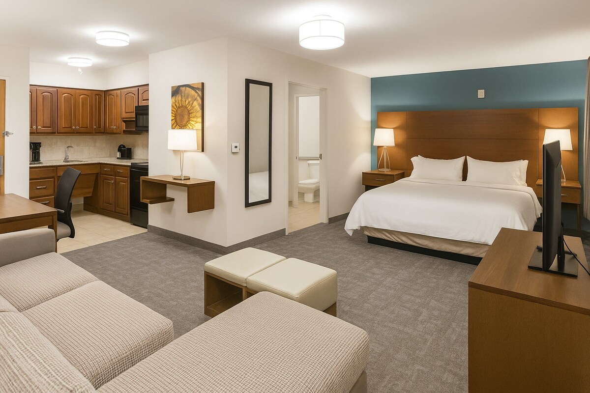 A hotel room with a bed, a desk, a chair, a sofa, a small table, a TV, and a bathroom visible through an open door.