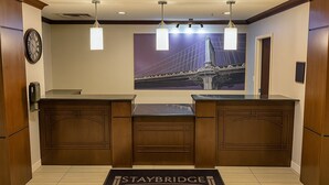 A reception area with a reception desk, a clock, pendant lights, and a framed picture of a bridge. - Staybridge Suites Elkhart North by IHG (Elkhart)