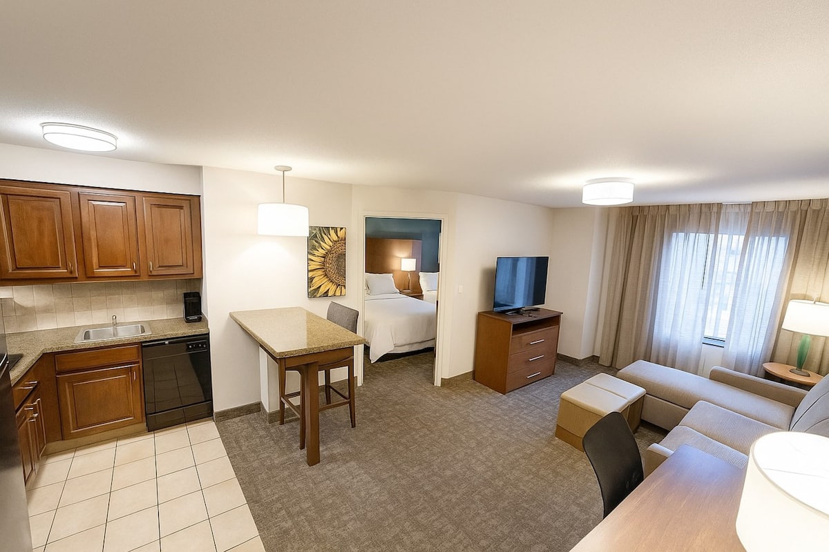 A compact hotel room with a kitchenette, a bed, a TV, a sofa, and a small dining area.