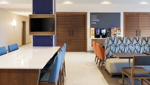 A modern hotel lobby with a long communal table, blue and orange seating, a coffee station, and a large wooden door. - Holiday Inn Express Lexington - Horse Park Area by IHG (Lexington)