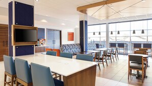 A modern dining area with a central white table surrounded by blue chairs, a flat-screen TV mounted on the wall, and large windows offering a view of the outside. - Holiday Inn Express Lexington - Horse Park Area by IHG (Lexington)