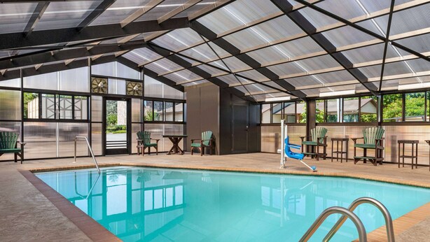 An indoor swimming pool with clear glass walls, surrounded by wooden deck chairs and tables. - Howard Johnson by Wyndham Pigeon Forge (Pigeon Forge)