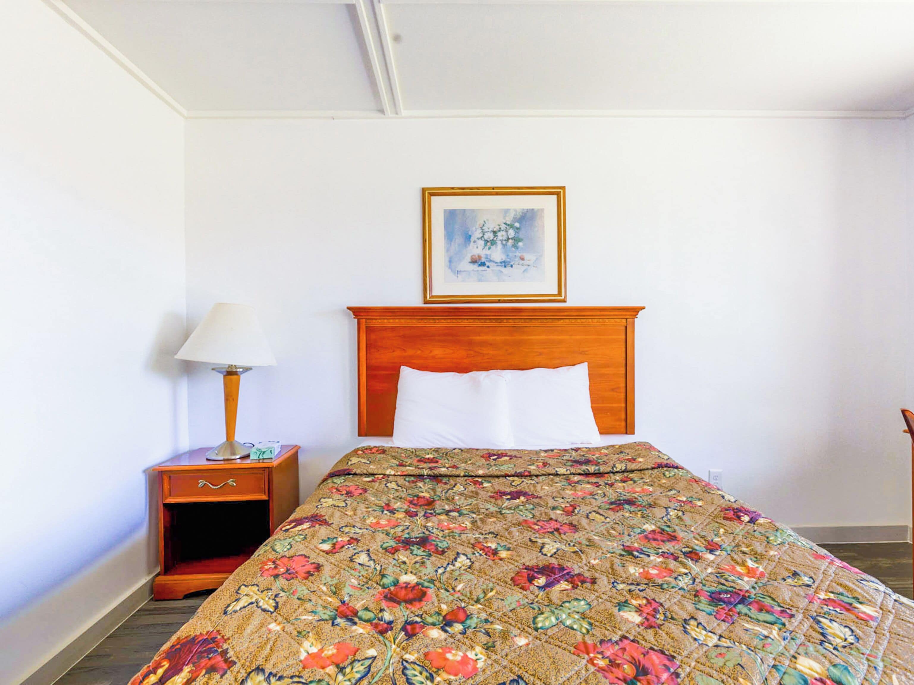 A hotel room with a wooden headboard, a floral bedspread, a nightstand with a lamp, and a framed picture on the wall.
