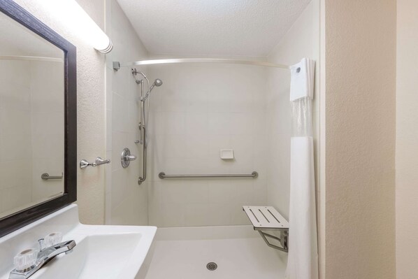 A small, clean bathroom with a white sink, a mirror, a shower with a grab bar, and a small shelf.
