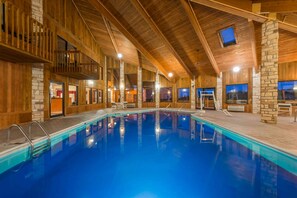 An indoor swimming pool with wooden walls and stone accents, surrounded by seating areas and a balcony.
