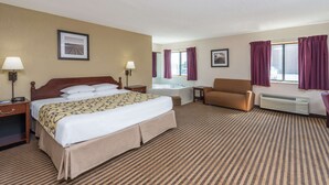A hotel room with a large bed, a chair, a desk, a desk lamp, a lamp, a window with curtains, a picture on the wall, and a bathroom with a bathtub and a mirror. - Baymont by Wyndham Metropolis (Metropolis)