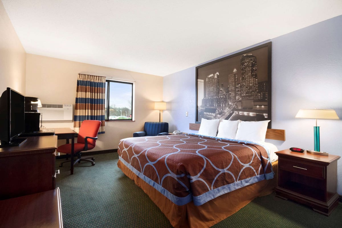 Habitación de hotel con una cama grande, un escritorio con computadora, una silla, una lámpara de escritorio y un ventanal con cortinas.