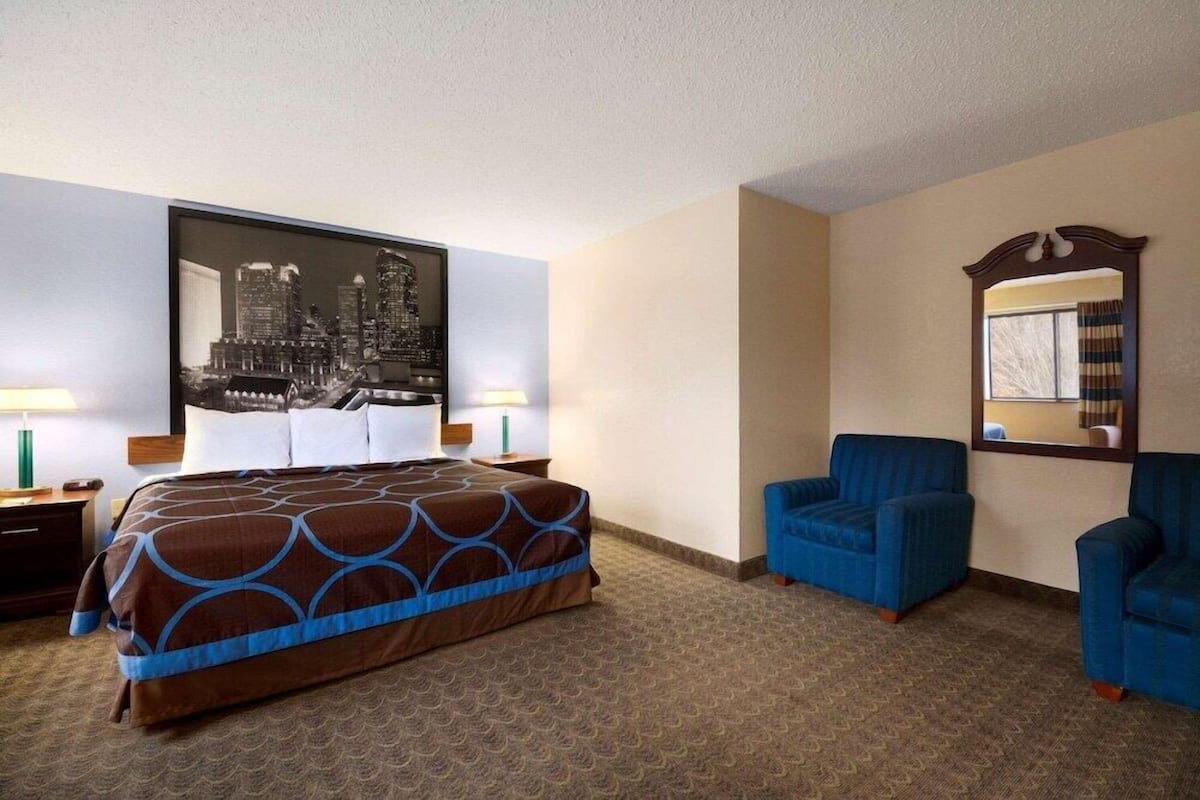 Una habitación de hotel con una cama, dos sillas, un espejo grande y un cuadro de un paisaje urbano en la pared.