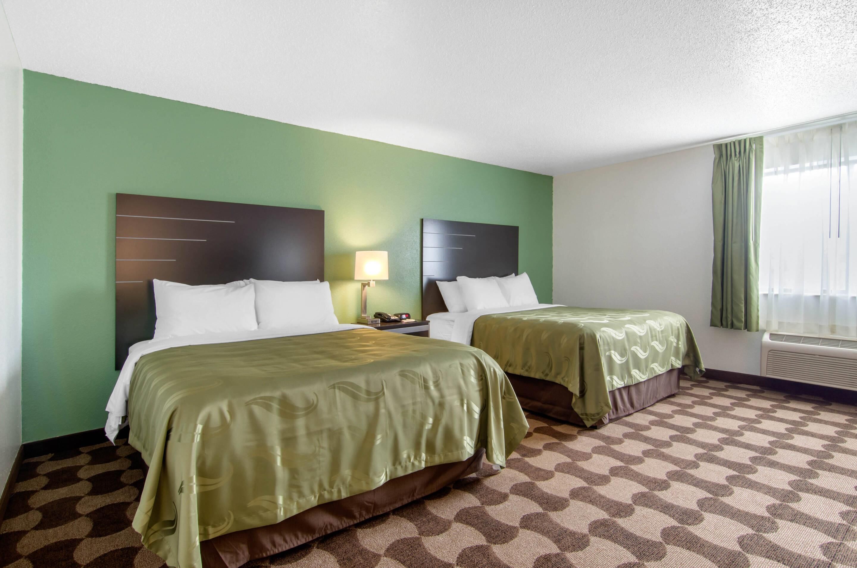 A hotel room with two beds, a green wall, a patterned carpet, and a window with curtains.