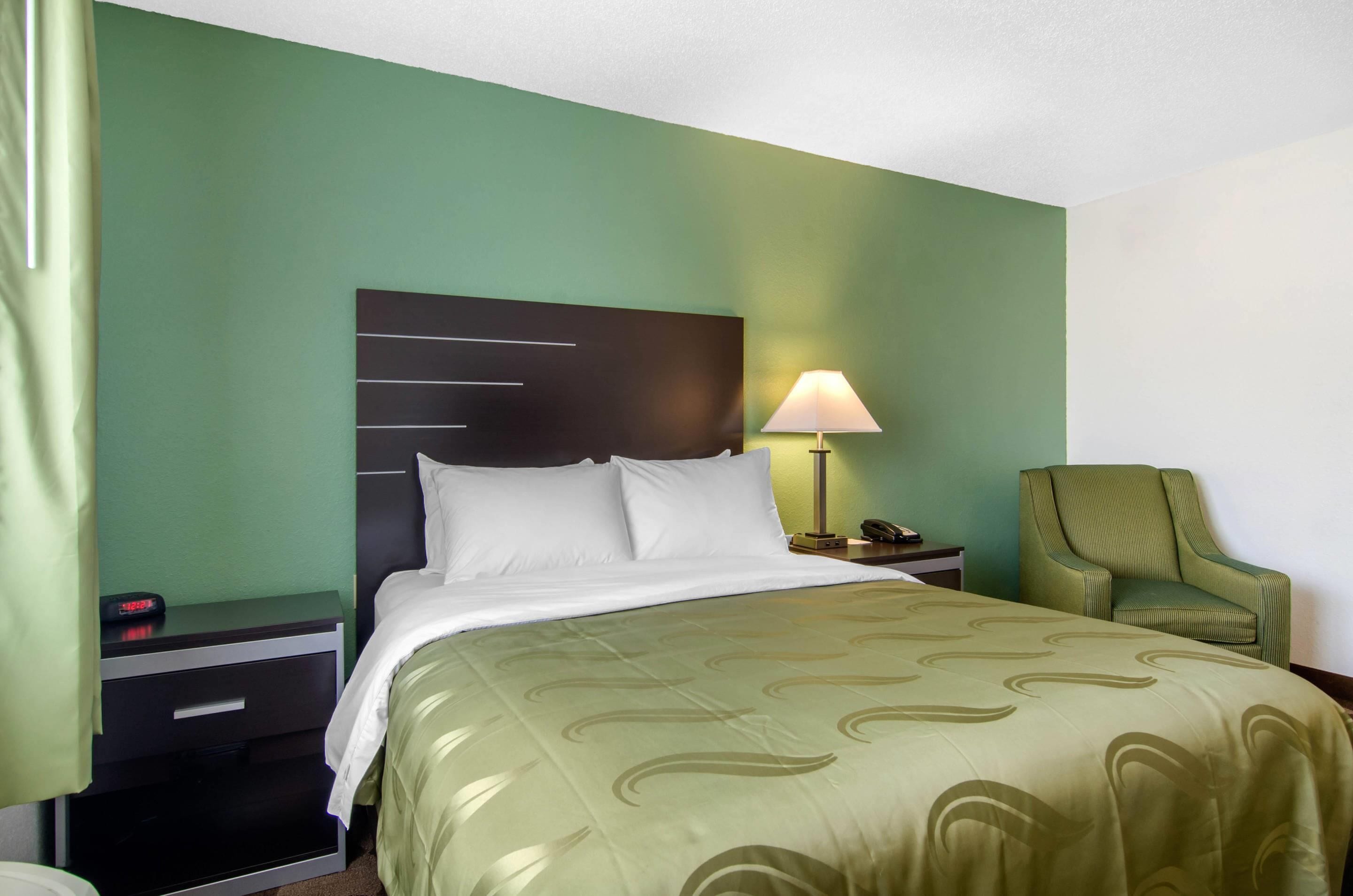 A hotel room with a bed, a nightstand with a lamp, a chair, and a wall clock.