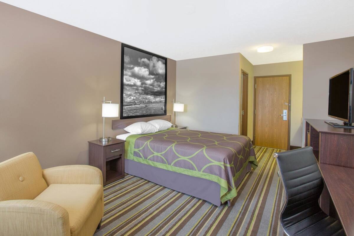 Une chambre d’hôtel comprenant un lit, un bureau avec une télévision, une chaise, une table de chevet et un tableau accroché au mur.
