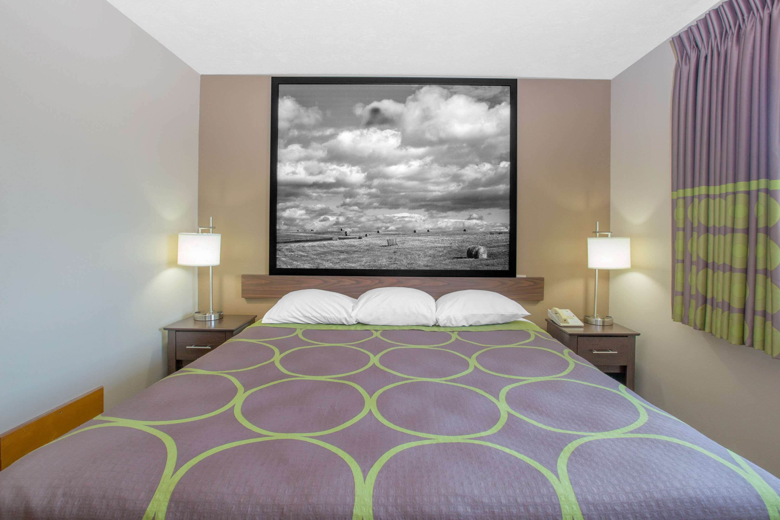 Una habitación de hotel con una cama, dos mesitas de noche, una lámpara de noche, un cuadro grande en la pared y una ventana con cortinas.