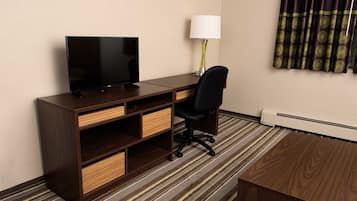 A hotel room with a wooden desk, a television, a lamp, a chair, and a carpeted floor.