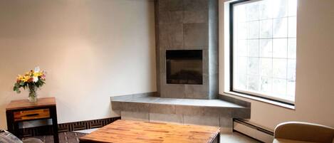 A living room with a fireplace, a large window, and a wooden coffee table.