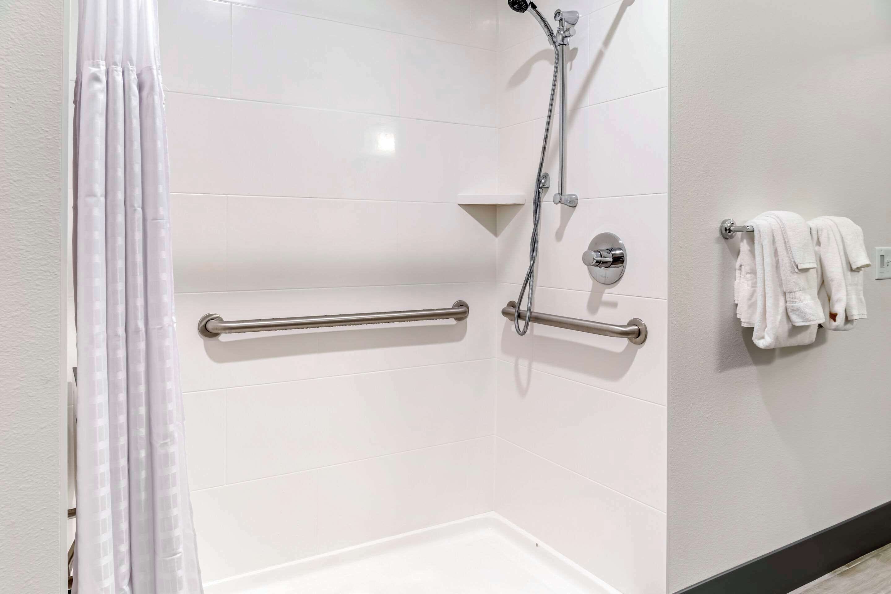 A modern bathroom with a shower, grab bars, a towel rack, and a white tiled wall.