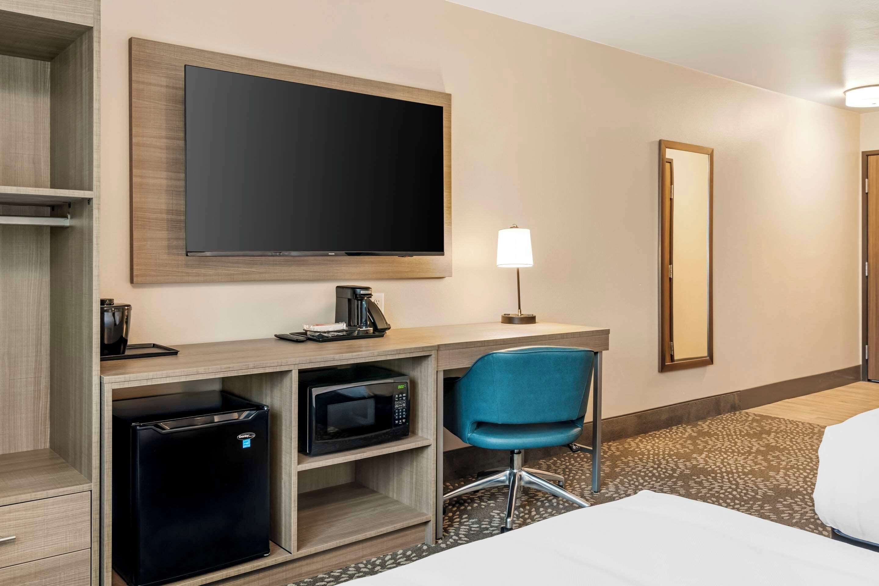 A hotel room with a flat-screen TV, a desk with a coffee maker, a microwave, a blue chair, and a bed.
