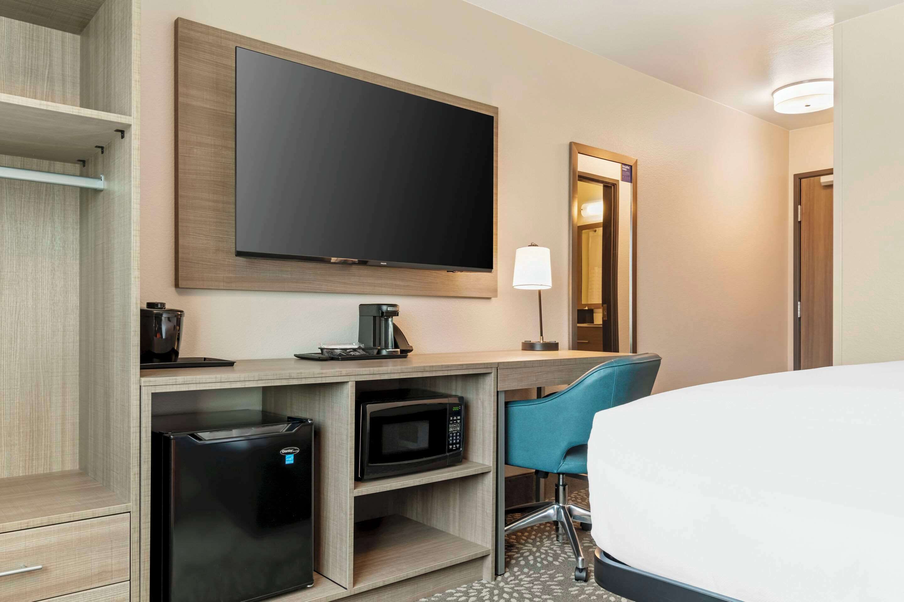 A hotel room with a bed, a desk with a computer, a microwave, a coffee maker, a TV, and a chair.
