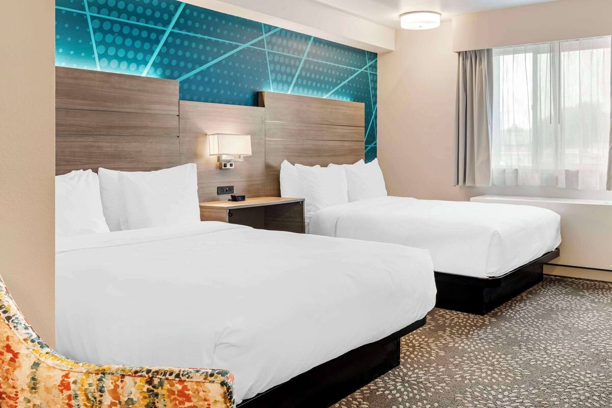 A hotel room with two beds, a nightstand, a lamp, and a window with curtains.