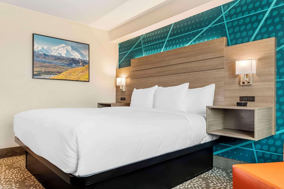 A hotel room with a large bed, wooden headboard, two bedside lamps, a nightstand, and a wall-mounted shelf.